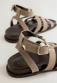 Braune Ledersandalen mit mehreren Riemen und goldfarbenen Schnallenverschlüssen vor schlichtem hellem Hintergrund.