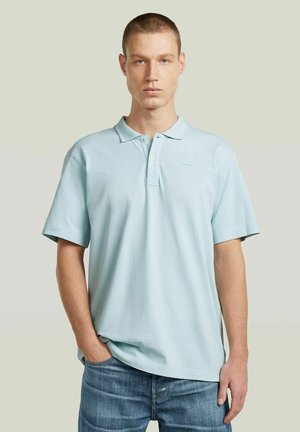 Junger Mann mit kurzen Haaren, der ein hellblaues Poloshirt und blaue Jeans trägt, steht mit einer Hand in der Tasche vor einem schlichten Hintergrund.