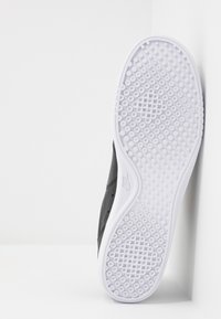 Svart sneaker med texturerad ovandel, vit gummisula med cirkulära greppmönster, lutad mot en vit bakgrund.