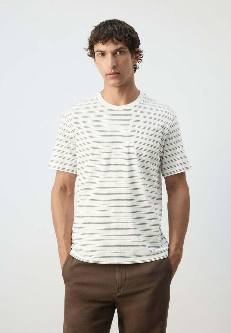 Jeune homme portant une chemise blanche à manches courtes avec de fines rayures horizontales noires et un pantalon marron, debout les mains dans les poches.