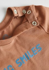 Nærbillede af en brun babydragt med metaltryknapper ved skulderen og blå tekst, der siger "STORE SMIL" på stoffet.