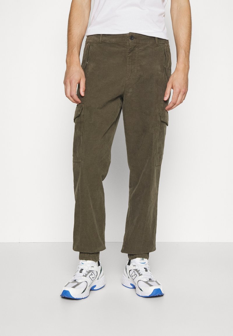 Homme portant un pantalon cargo vert olive avec des poignets élastiques et des baskets blanches avec des accents bleus, debout devant un fond blanc uni.