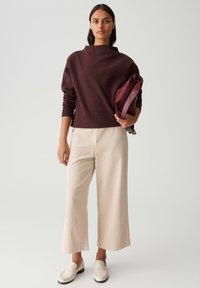 Vrouw die staat, gekleed in een bordeauxrode lange mouwen top, crème wijde broek, witte loafers, en een bijpassende bordeauxrode tas onder haar arm houdt.