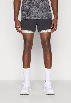 Patroon sporttop met grijs en witte ontwerpen, gecombineerd met zwarte shorts over gestreepte compressieshorts. Witte sportkousen en schoenen.