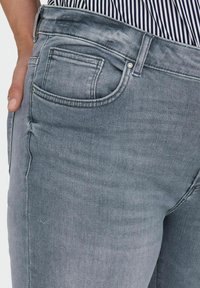 Vaqueros de denim gris claro con un diseño ajustado, que cuentan con un bolsillo trasero y costuras en tono. La textura es suave con un ligero brillo.