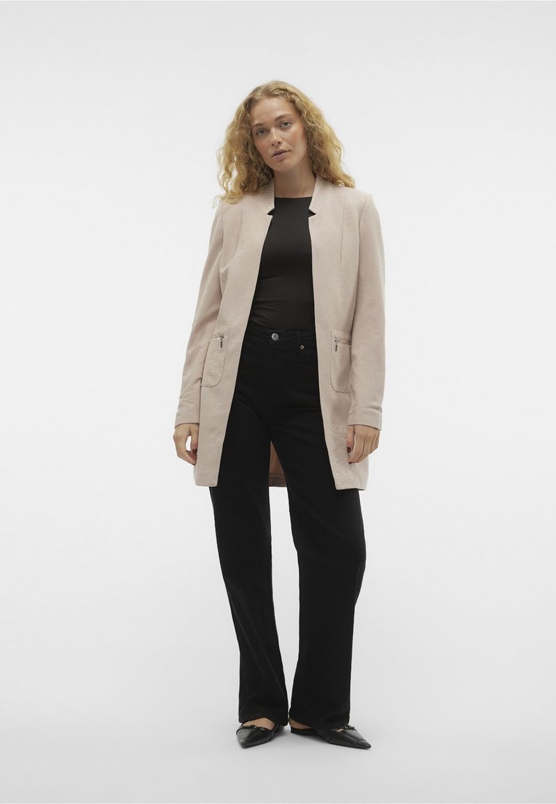 Blazer ajustado de color rosa claro con un acabado texturizado, con abertura frontal, dos bolsillos delanteros, y combinado con pantalones de pierna ancha negros y zapatos planos de punta.
