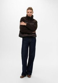 Frau steht mit verschränkten Armen, trägt eine dunkelbraune Fleecejacke, dunkelblaue Jeans und braune Schuhe vor einem schlichten weißen Hintergrund.