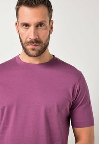 Homme aux cheveux courts bruns et à la barbe, portant un t-shirt violet uni à col rond, sur un fond blanc uni.