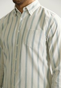 Mężczyzna ubrany w koszulę z długim rękawem, zapinaną na guziki, w pionowe jasnoniebieskie i kremowe paski, z kieszonką na piersi, na jednolitym tle.