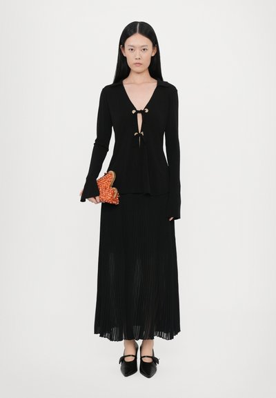 Black long-sleeve top with a deep V-neck and gold accents, paired with a pleated black skirt and pointed black shoes, holding an orange textured clutch.