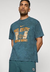 Blå tie-dye t-shirt lavet af bomuld med orange grafisk tekst, der siger "Nike Athletic Department." Har en rund halsudskæring og korte ærmer.