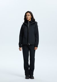 Veste noire isolée avec une fermeture éclair à l'avant, deux poches poitrine et une capuche réglable, portée avec un pantalon noir assorti et des bottes robustes.