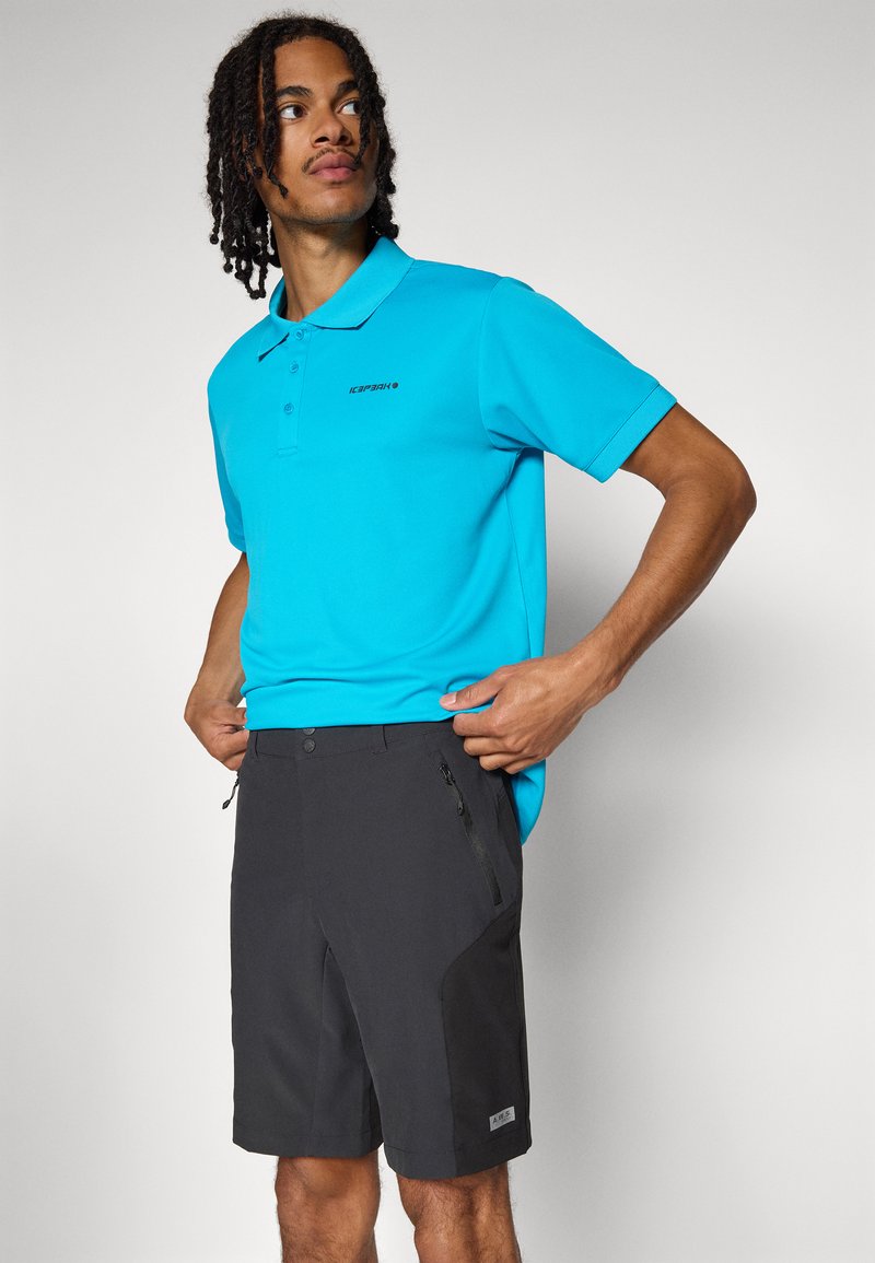 Türkises Poloshirt mit drei Knöpfen und Logo, kombiniert mit schwarzen Shorts mit Reißverschluss-Taschen und kontrastierendem Seiteneinsatz.