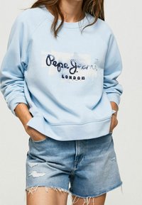 Persona che indossa un felpa leggera blu di Pepe Jeans London e pantaloni shorts in denim strappati, con le mani infilate nelle tasche, contro uno sfondo semplice.