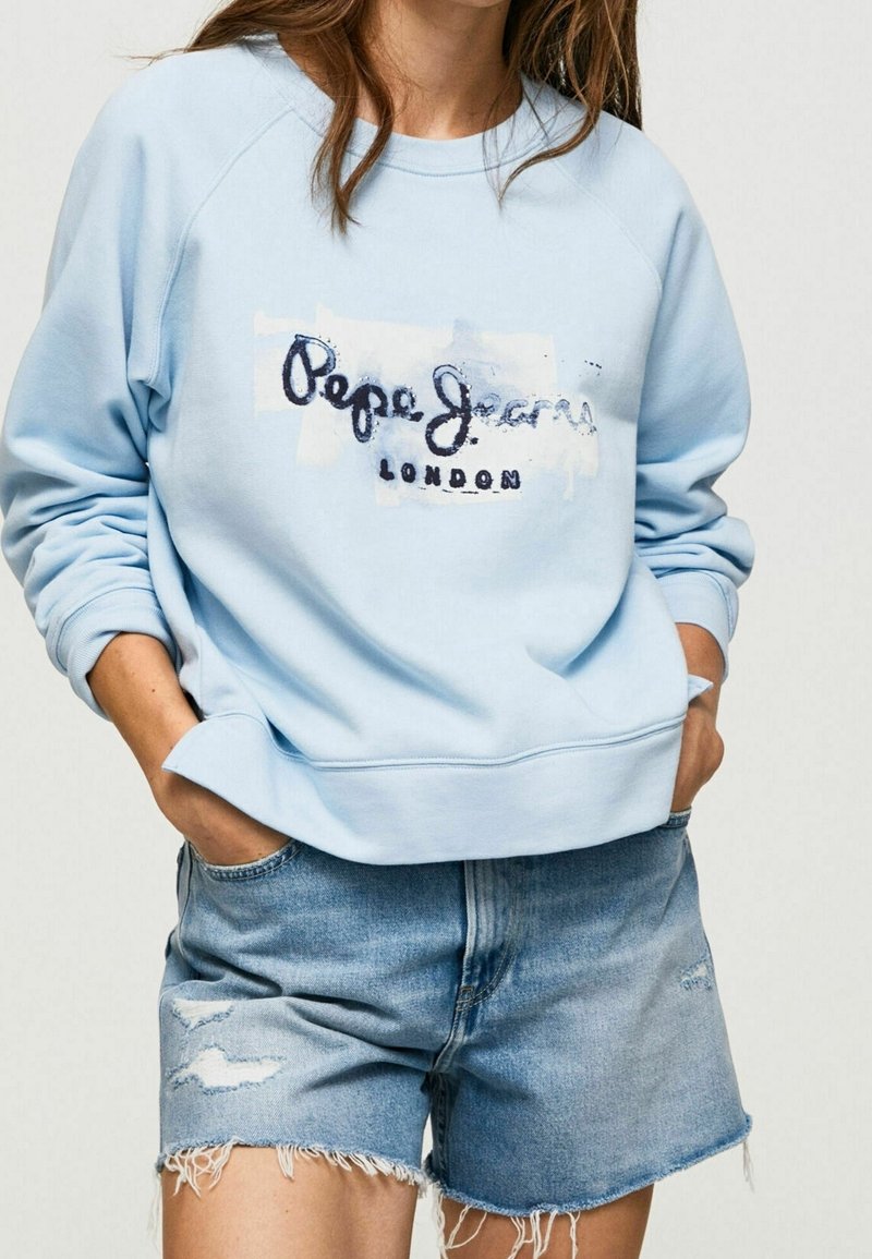 Persona che indossa un felpa leggera blu di Pepe Jeans London e pantaloni shorts in denim strappati, con le mani infilate nelle tasche, contro uno sfondo semplice.