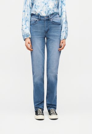 Person iført lyseblå blomstret bluse, lige-ben mediumblå jeans og sorte og hvide sneakers, stående mod en hvid baggrund.