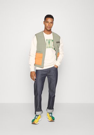 Chaleco acolchado verde claro con bolsillos naranjas sobre un suéter blanco con texto verde, combinado con jeans oscuros enrollados y zapatillas multicolores.