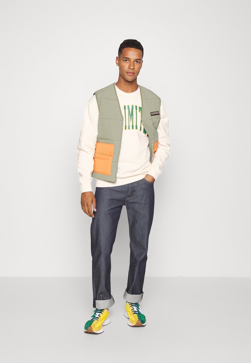 Gilet matelassé vert clair avec poches orange sur un pull blanc avec du texte vert, associé à un jean bleu marine retroussé et des baskets multicolores.