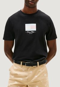 Czarna bawełniana koszulka z prostokątnym wzorem logo w jasnoniebieskim i czerwonym kolorze, z napisem "Tommy Hilfiger" oraz "EST. 1985" poniżej.