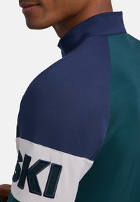 Mężczyzna w kurtce z wysokim kołnierzem w kolorach granatowym, morskim i białym, z haftem "SISKI" na rękawie.