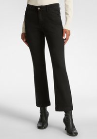 Vaqueros acampanados negros de denim, con cierre frontal de botón, dos bolsillos delanteros y una textura limpia y suave. Combinados con botas negras hasta el tobillo.