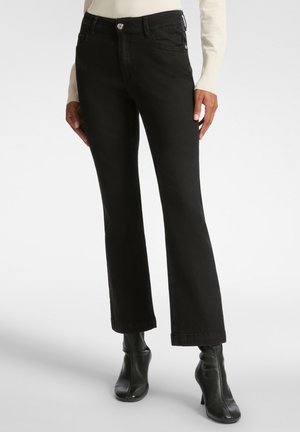 Vaqueros acampanados negros de denim, con cierre frontal de botón, dos bolsillos delanteros y una textura limpia y suave. Combinados con botas negras hasta el tobillo.