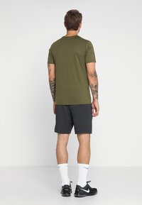 Man med tatueringar klädd i en olivgrön t-shirt, mörkgrå shorts, vita Nike-strumpor och svarta Nike Air Max sneakers, stående vänd mot en vit vägg.