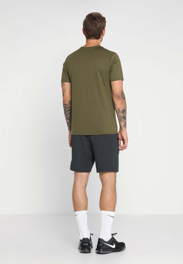 Man med tatueringar klädd i en olivgrön t-shirt, mörkgrå shorts, vita Nike-strumpor och svarta Nike Air Max sneakers, stående vänd mot en vit vägg.