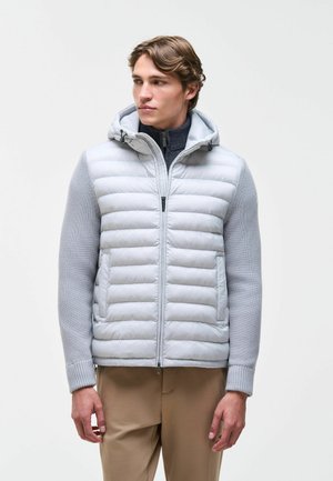 Junger Mann, der eine hellgraue wattierte Jacke mit Strickärmeln, beige Hose trägt und seitlich gegen einen einfarbigen Hintergrund blickt.