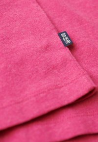 T-shirt rosa a maniche corte in cotone, con una consistenza morbida e un'etichetta sull'orlo che mostra testo nero su uno sfondo blu.