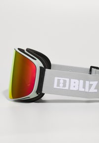 Gogle narciarskie z zakrzywioną, wielokolorową soczewką przechodzącą od czerwonego do żółtego. Szary, regulowany pasek z białym napisem "BLIZ". Miękka piankowa wyściółka.