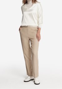 Pantalons beige ajustés avec une coupe slim et des plis sur le devant, associés à un top en soie crème à col en V et des mocassins blancs. Texture lisse et design minimaliste.