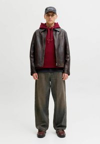 Ruskea nahkatakki burgundinvärisen hupparin päällä, jossa on narut, yhdistettynä leveisiin vihreäruskeisiin farkkuihin ja tummiin kenkiin, täydentäen asua musta hattu.