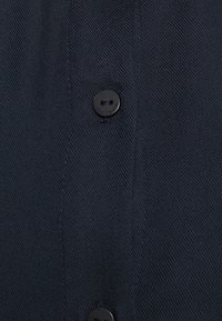 Svart knappad skjorta i texturerad tyg, med en centrerad knäppning och släta knappar. Närbild som framhäver detaljerna i sömnaden.