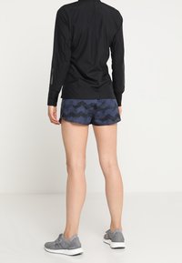 Chemise noire à manches longues, shorts bleu marine à motif zigzag, et chaussures de sport grises, présentant une tenue sportive adaptée à une activité physique.