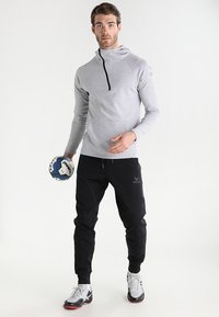 Pull-over de sport gris avec un col montant et une fermeture éclair noire, associé à un pantalon de jogging noir avec un logo en relief. Tenant une balle de handball bleu et blanc.