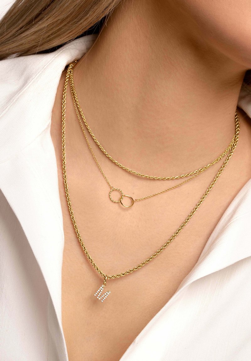 Collane a strati in oro con design a catena attorcigliata. Una presenta un piccolo dettaglio circolare, l'altra ha un ciondolo con la "M" in strass.