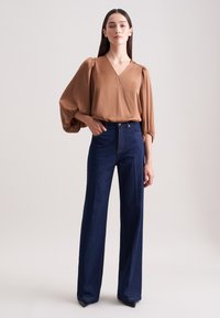Brun silke bluse med puffærmer, parret med højttaljede mørkeblå denim flared bukser. Glat tekstur, v-udskæring og skræddersyet pasform.