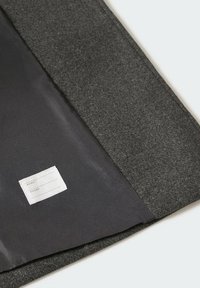 Šedá plstěná obálka s černým textilním vnitřkem, obsahující malý štítek pro jméno a telefon. Jednoduchý, nenápadný design a textura.