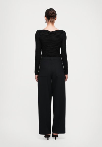 Woman with hair in bun wearing a fitted textured black long-sleeve top, wide-leg black pants, and black heeled shoes, standing facing away.