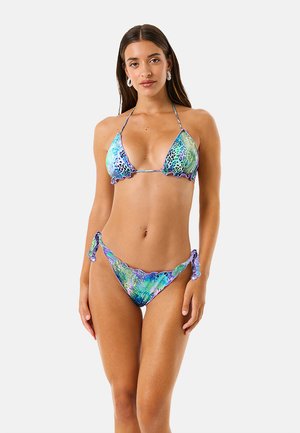 Donna che indossa un bikini a balze con stampa animalier blu e verde, con slip con laccetti ai lati, in piedi su sfondo bianco uniforme.