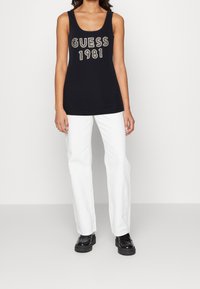 Donna che indossa un top senza maniche nero con la scritta "GUESS 1981", pantaloni bianchi a gamba dritta e scarpe con zeppa nere su uno sfondo neutro.