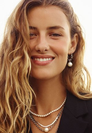Zilveren en parel sieraden gelaagd rond de nek; krullend, golvend haar; zwarte blazer; zachte focus achtergrond.