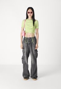 Ljus limegrön ribbad crop top med dragsnören, tillsammans med grå cargobyxor med sidofickor och en avslappnad passform. Rosa handväska och solglasögon.