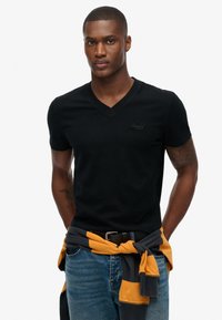 Sort kortærmet V-hals t-shirt med et diskret logo, parret med blå jeans og en grå-gul stribet sweater bundet om taljen.