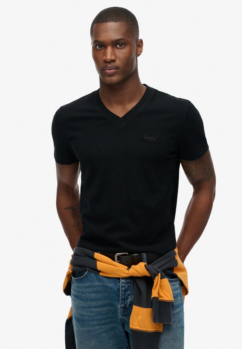 Sort kortærmet V-hals t-shirt med et diskret logo, parret med blå jeans og en grå-gul stribet sweater bundet om taljen.