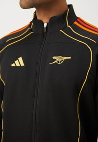 Veste noire à fermeture éclair avant avec des accents dorés, présentant des rayures rouges et orange sur les épaules et un logo d'Arsenal brodé en doré.