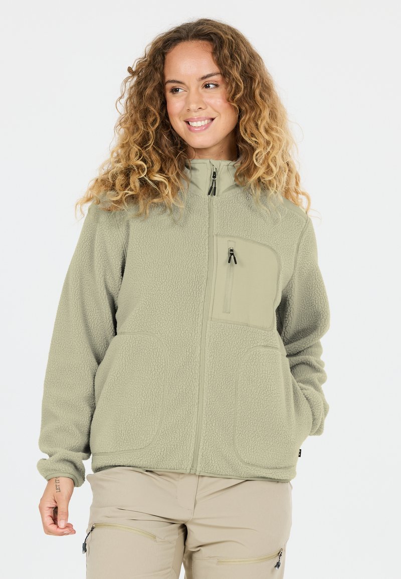 Vrouw met krullend haar die een lichtgroene fleecejas met rits en beige broek draagt, glimlachend en zijwaarts kijkend tegen een witte achtergrond.