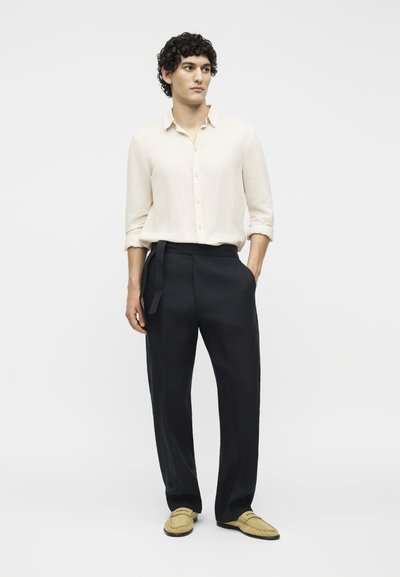 Άνδρας με κατσαρά μαλλιά που φοράει κρεμ μακρυμάνικο πουκάμισο, μαύρο παντελόνι με πλαϊνή ζώνη και μπεζ μοκασίνια, στέκεται με τα χέρια στις τσέπες.