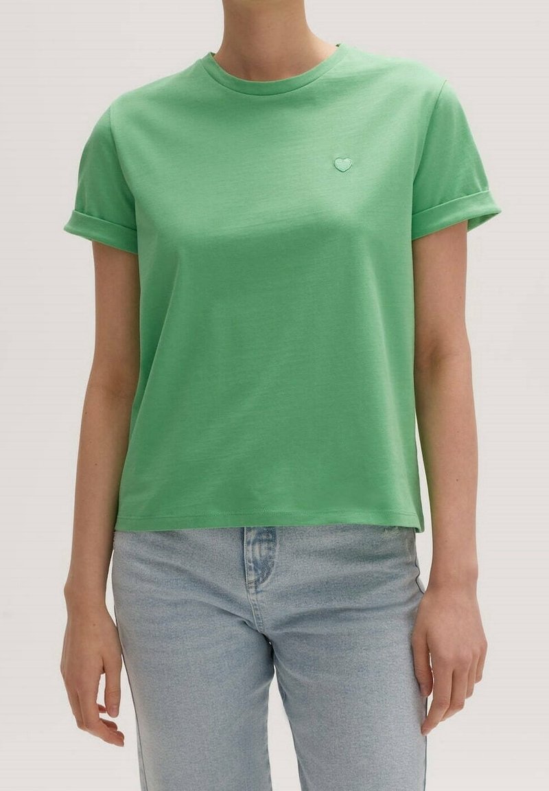 Groene T-shirt met korte mouwen van zachte katoen, voorzien van een ronde halslijn en een klein hartembleem op de linkerkant van de borst. Gecombineerd met lichtblauwe jeans.